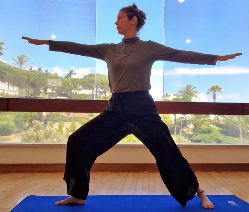 Netta in a yoga warrior pose in a studio overlooking trees
